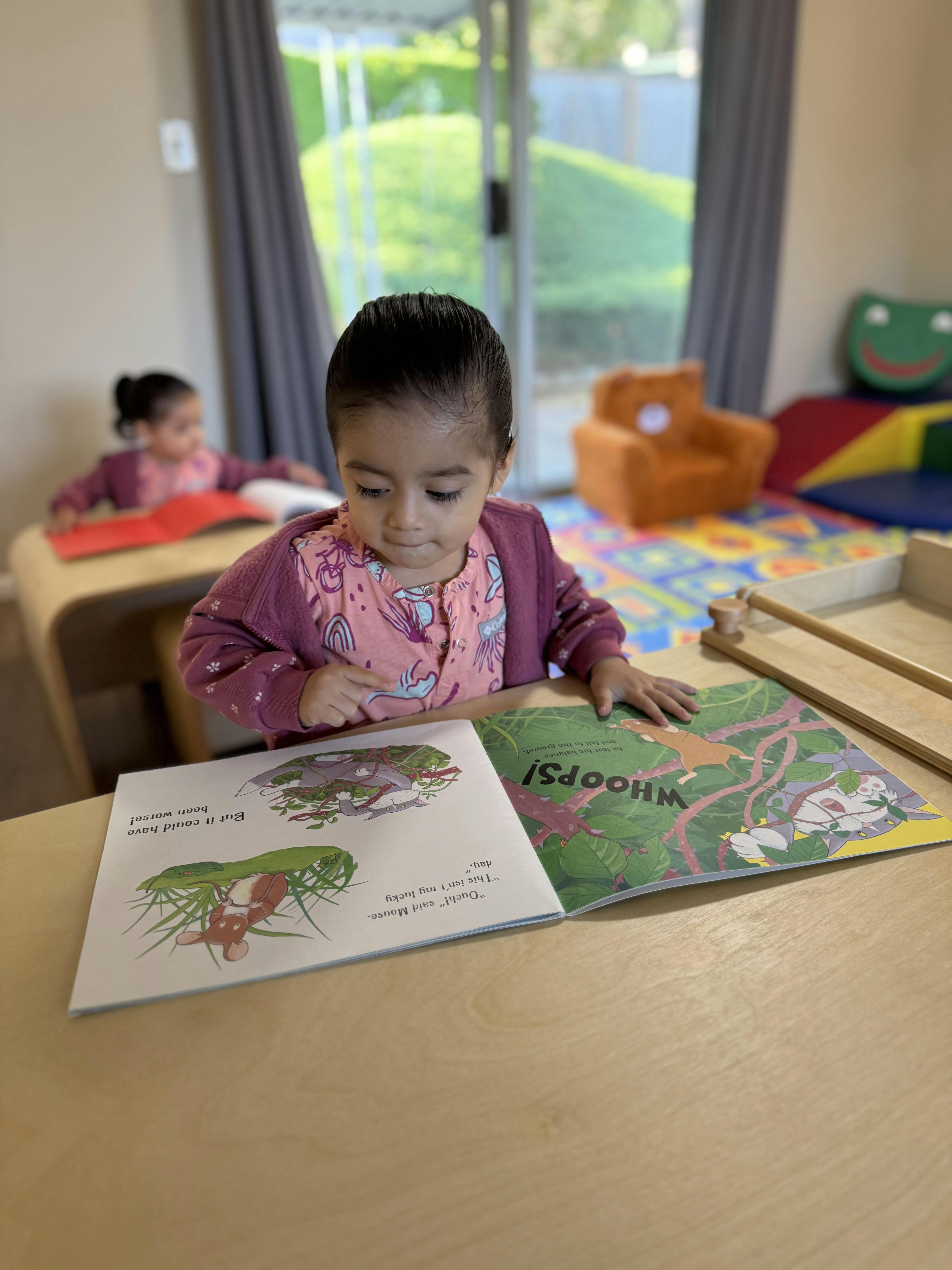 Child reading a book at a table, another child in the background