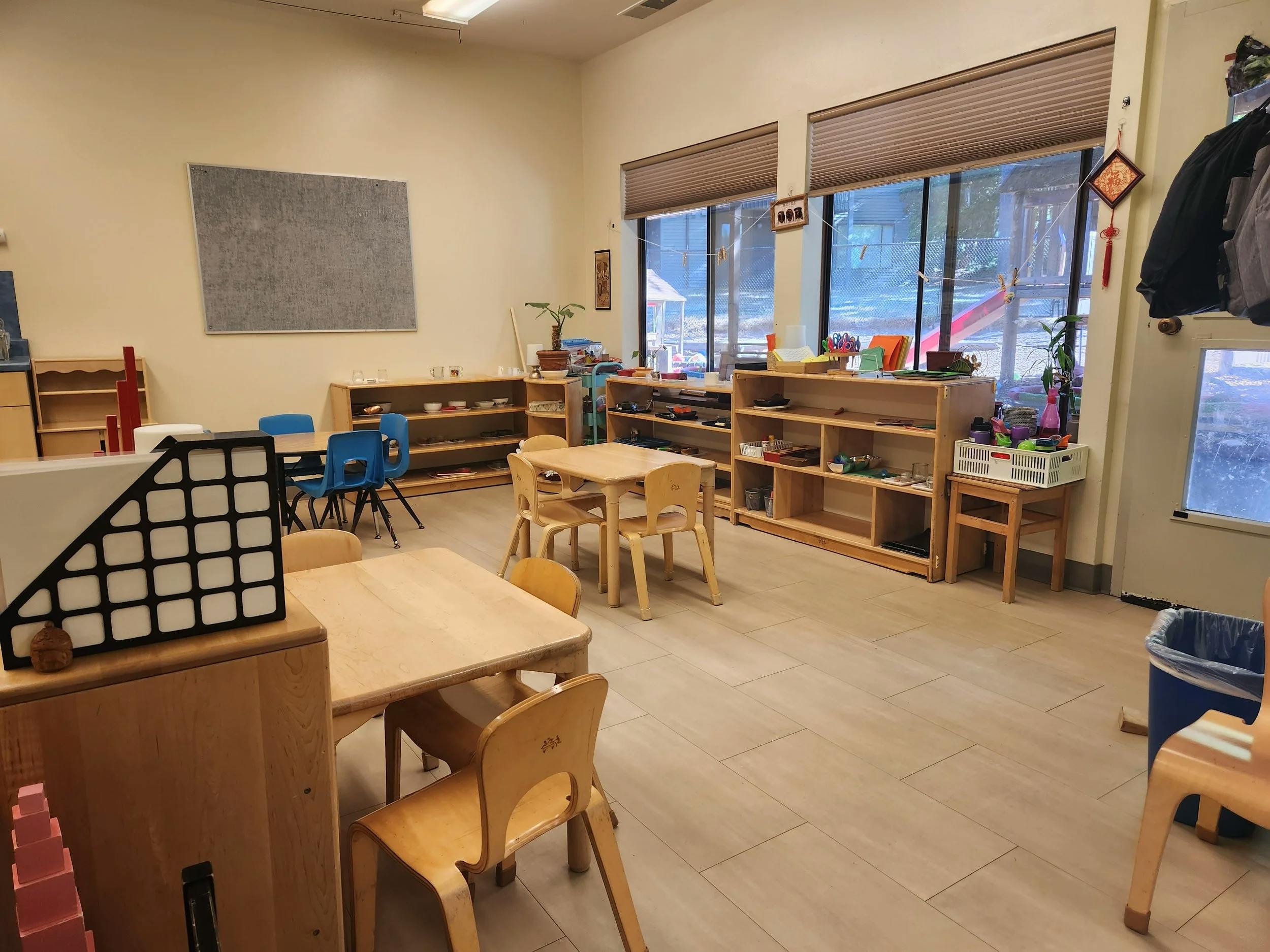 Classroom with small wooden tables and chairs, shelves with learning materials