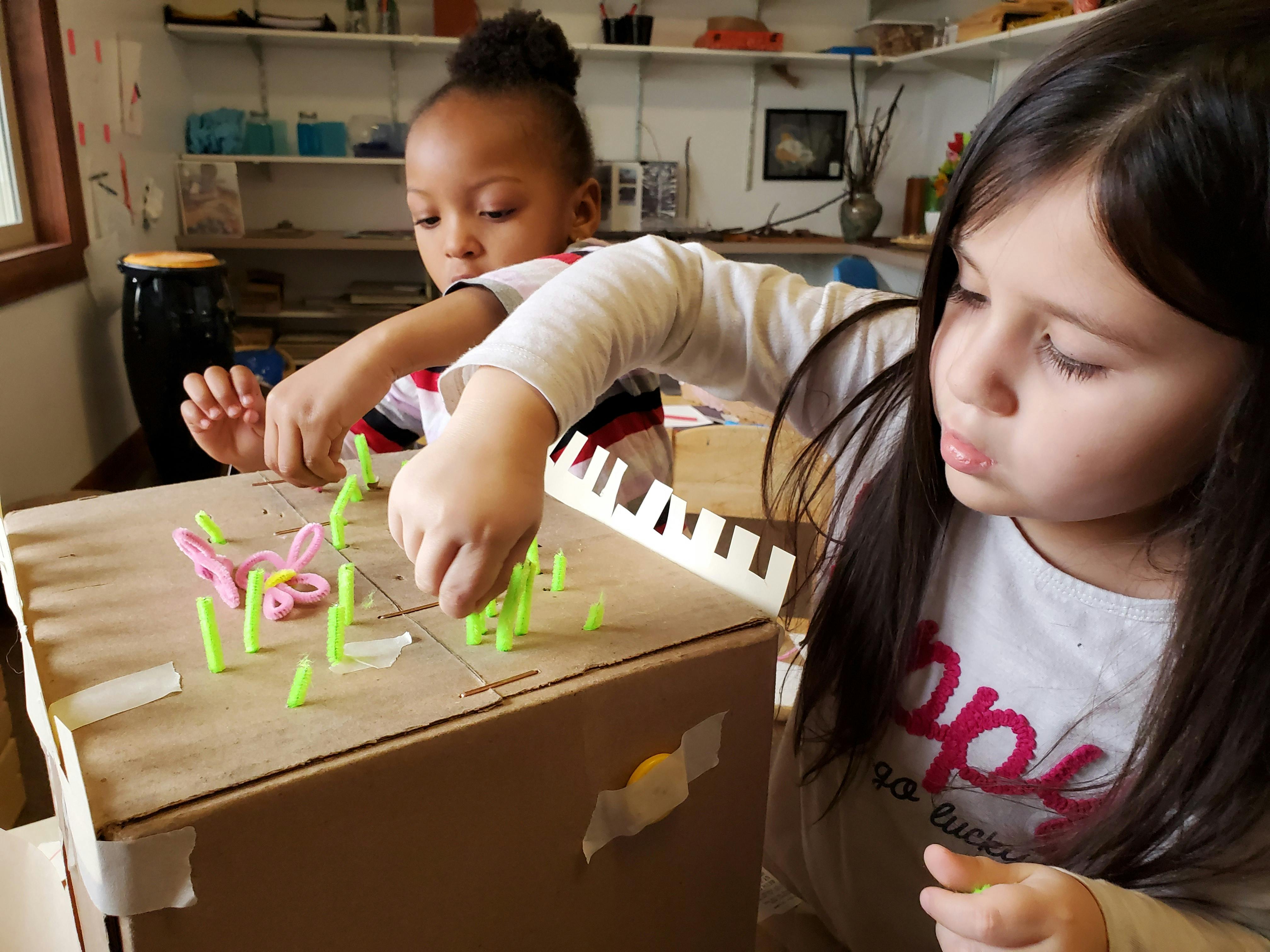 Two children playing with pipe cleaners on a cardboard box