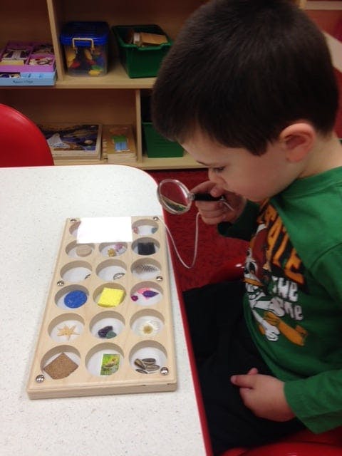 Child looking through a magnifying glass at items in a wooden tray