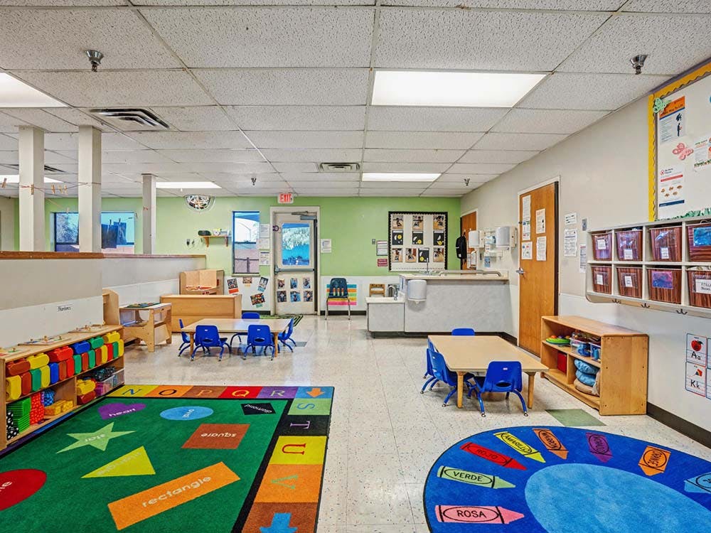 An empty daycare classroom with small tables and chairs, colorful rugs, and play areas