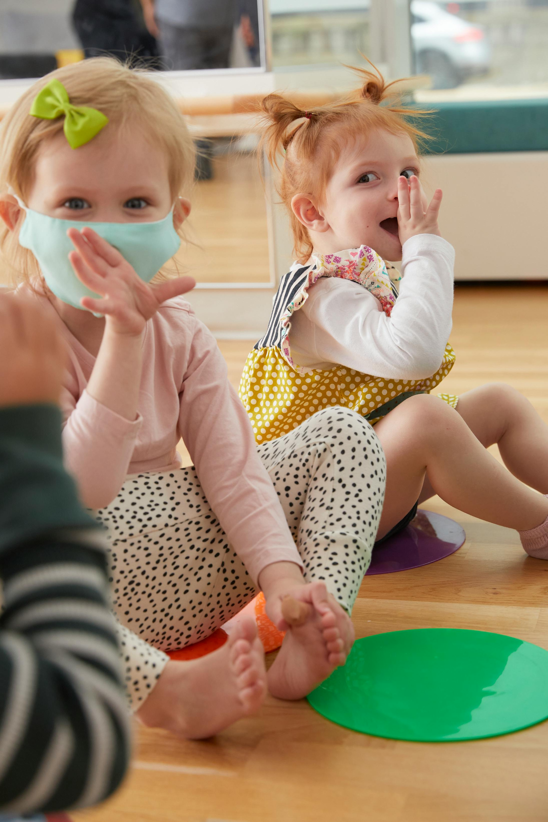 Two children sitting on a wooden floor, one wearing a face mask