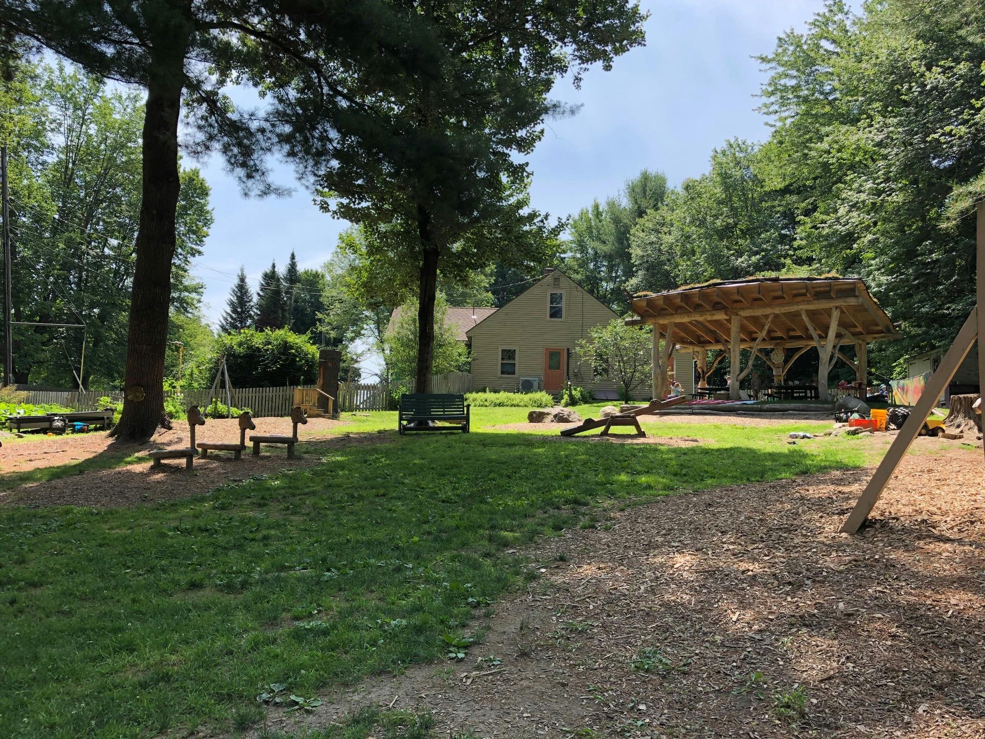 Outdoor play area with grass, trees, a wooden structure, and a house in the background