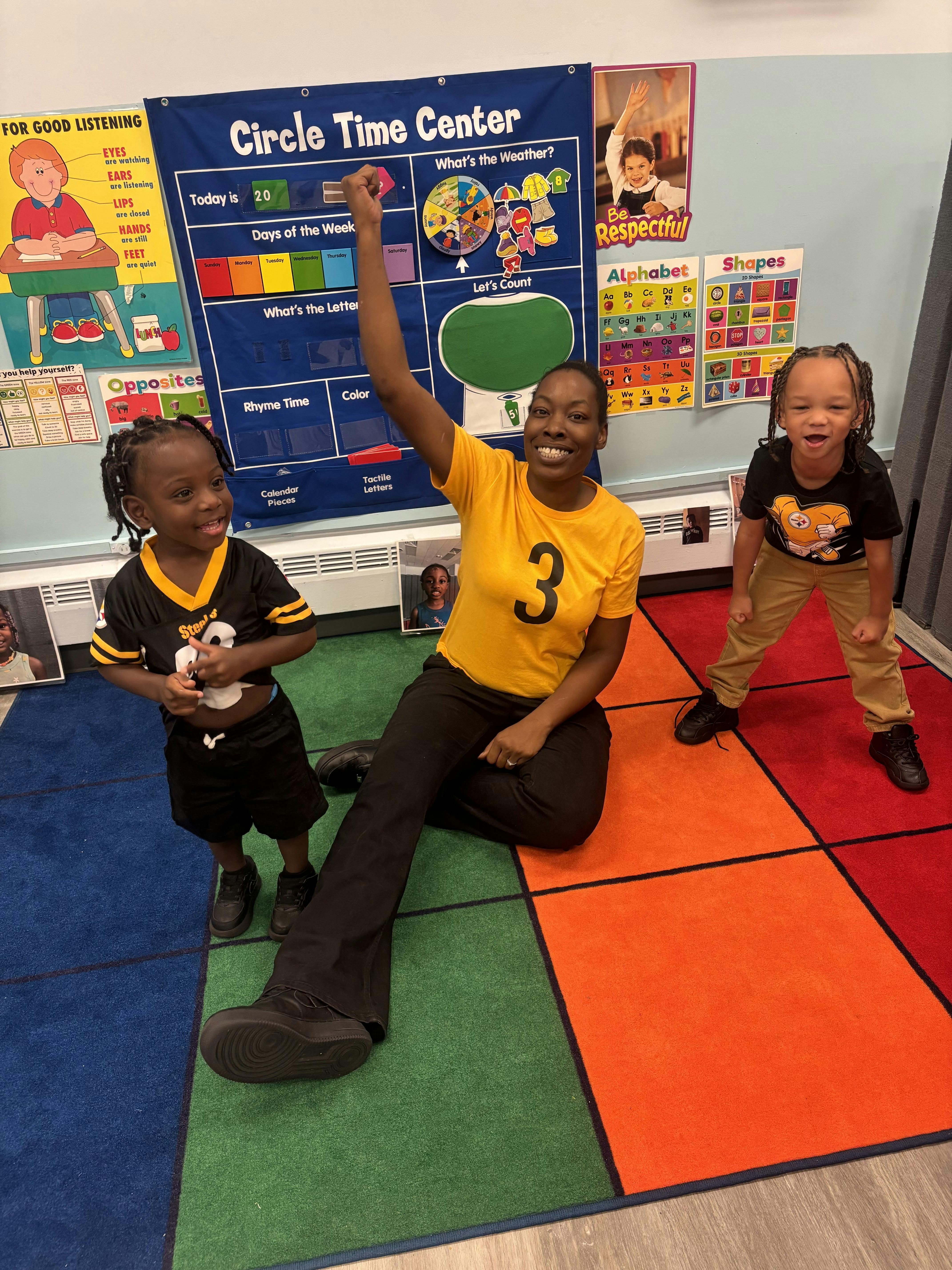 A person and two children in a classroom in front of a circle time chart