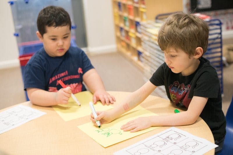 Two children drawing on paper at a table in a classroom