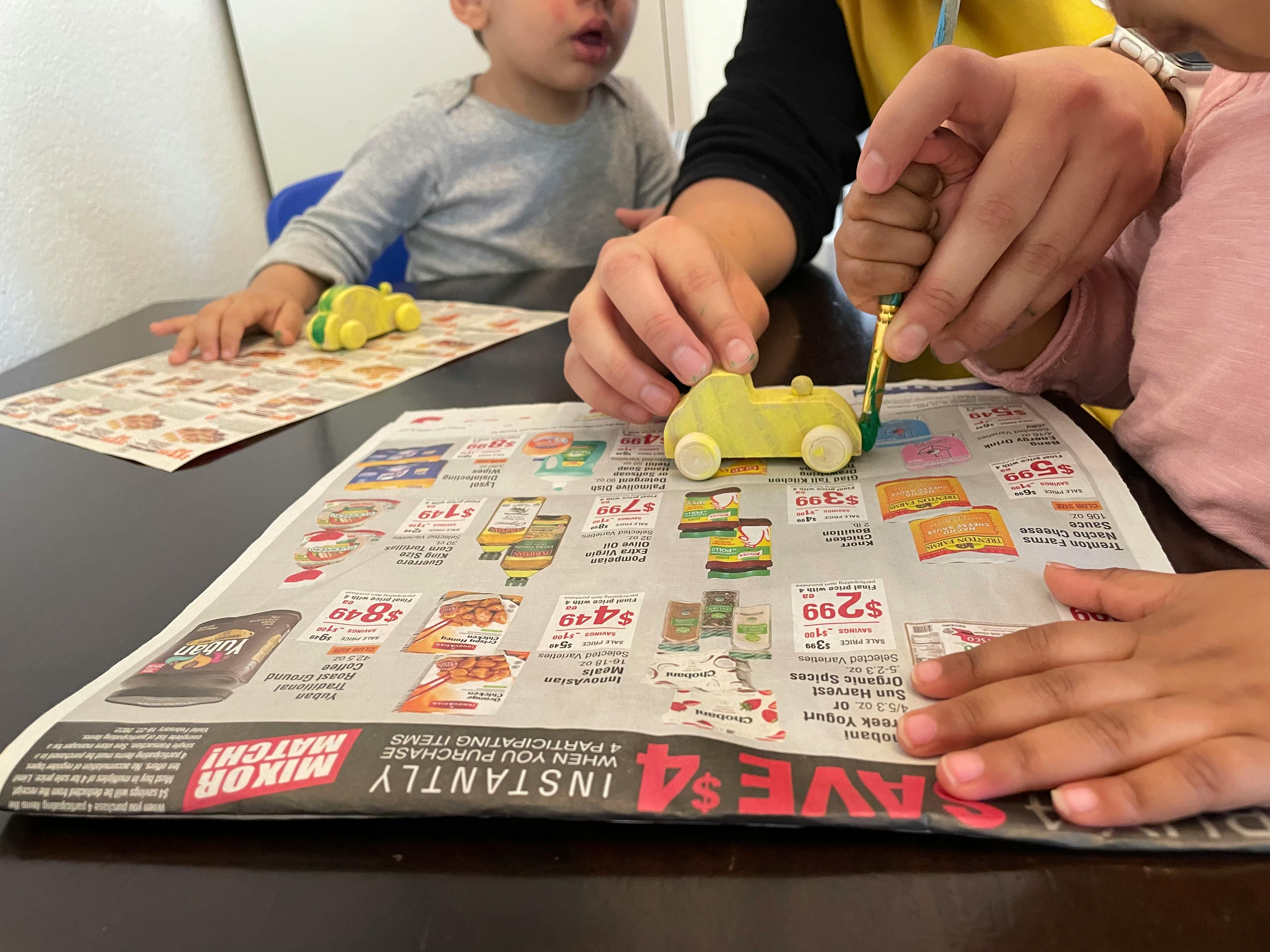 Person painting a small wooden car on a newspaper-covered table