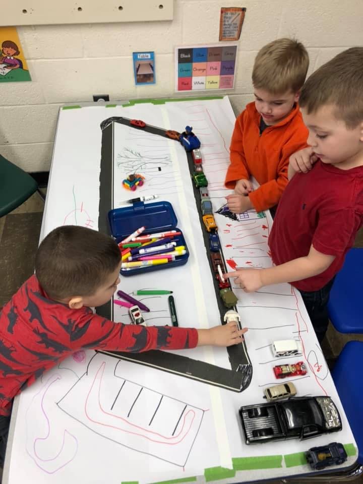 Three children playing with toy cars on a drawn road on a table