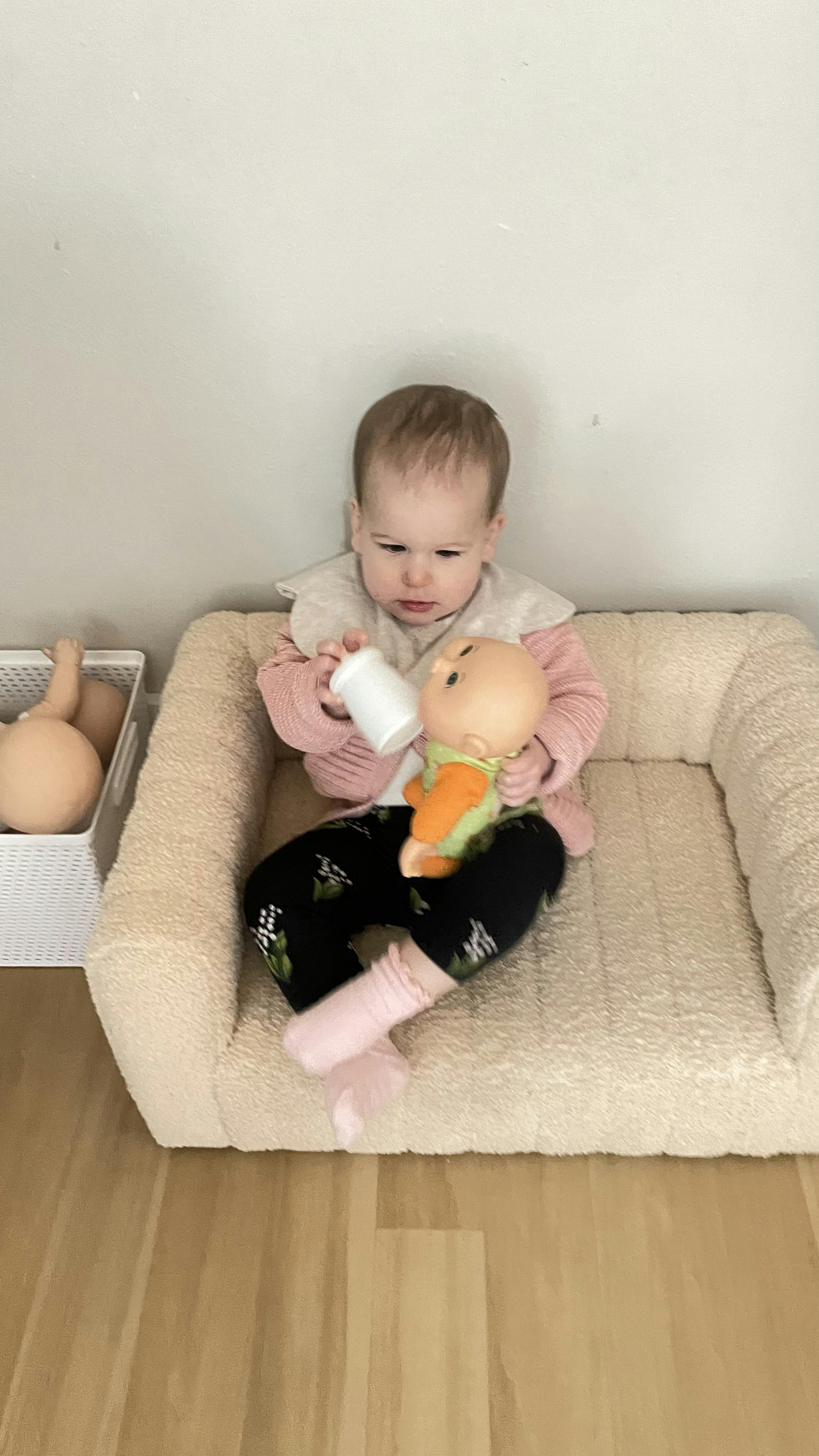 Child sitting in a cushioned pet bed, feeding a doll with a bottle