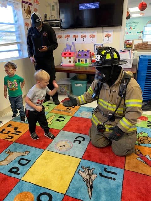 Firefighter in gear kneeling on a rug with a child in a classroom