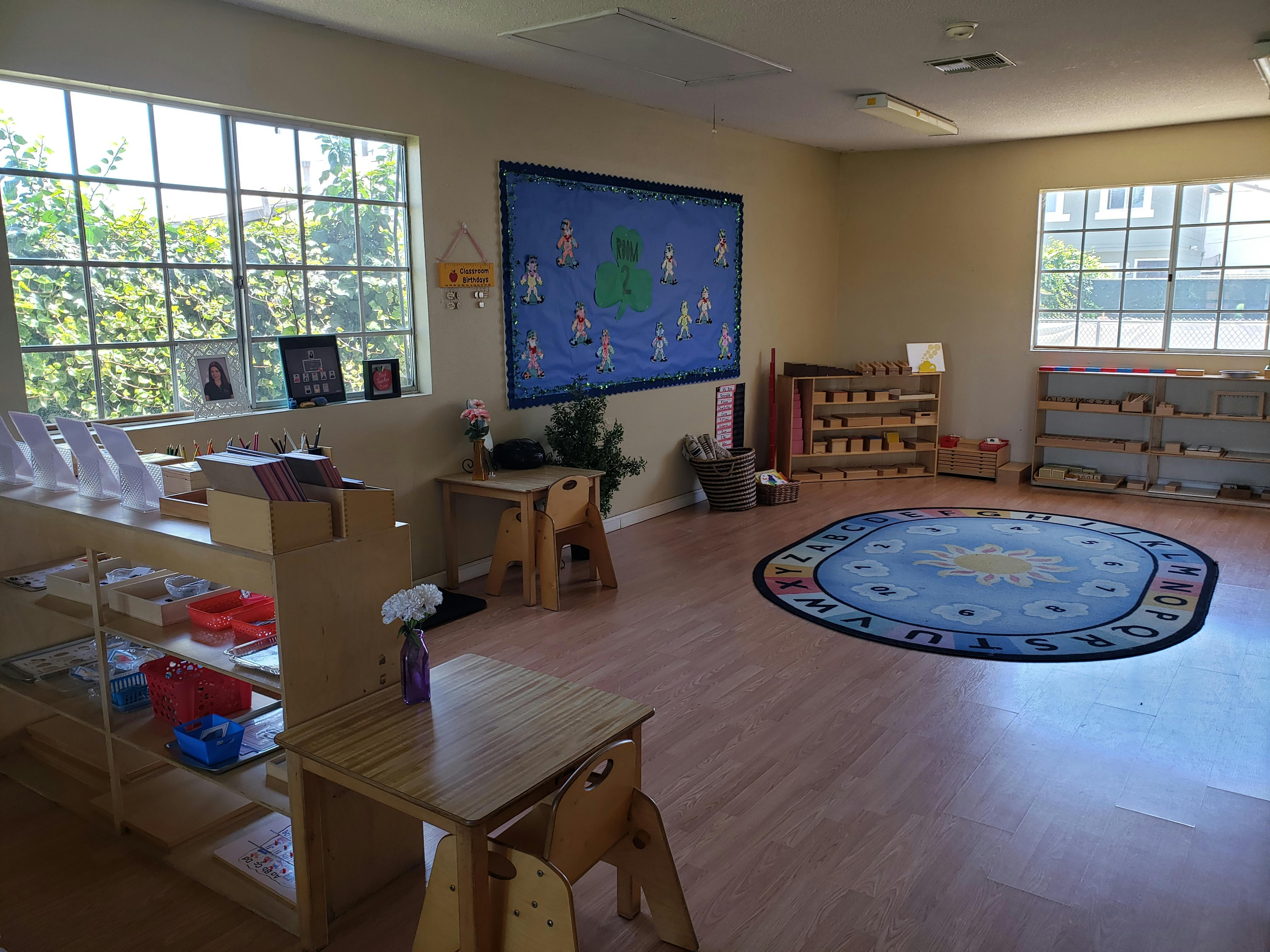 Classroom with small wooden tables and chairs, shelves, and a blue rug