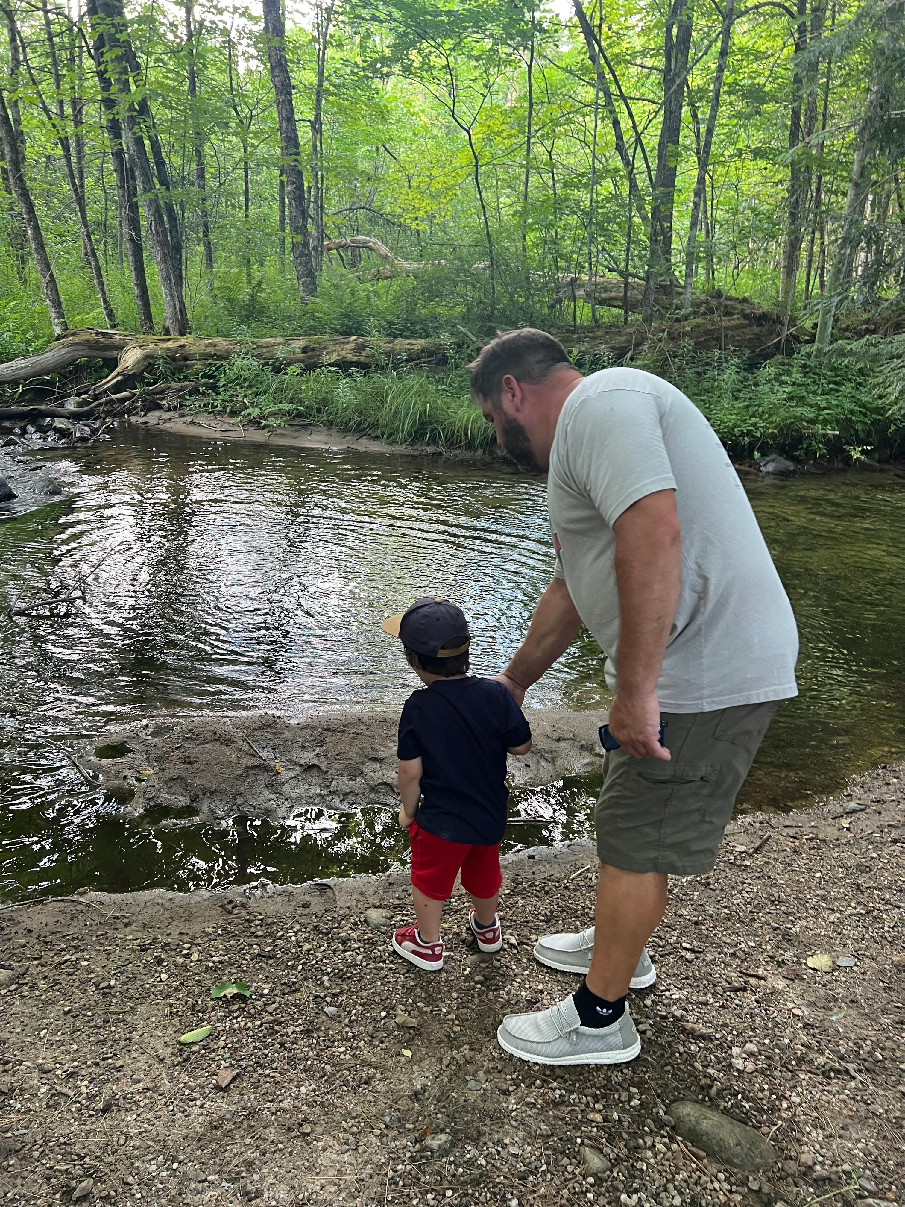 Person and child standing by a stream in a wooded area