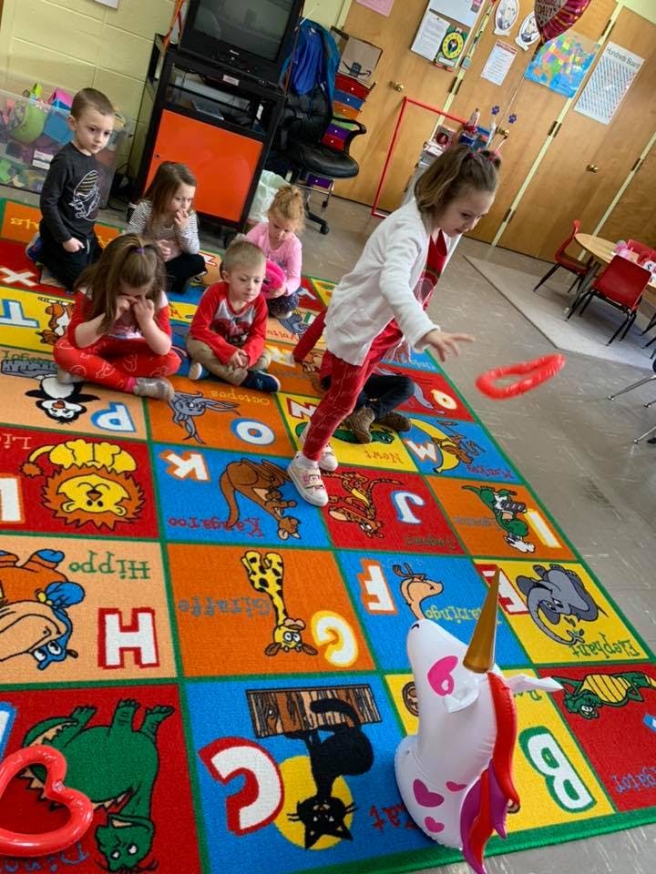 Child throwing a red ring toward a unicorn toy on a colorful alphabet rug with other children