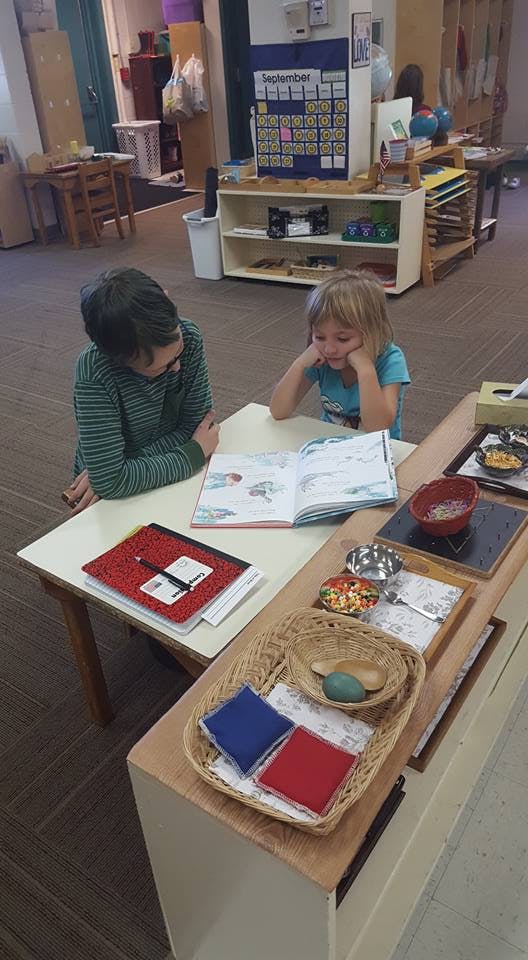 Two children looking at an open book on a table in a classroom
