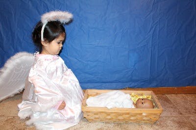 Child in angel costume looking at a baby doll in a basket