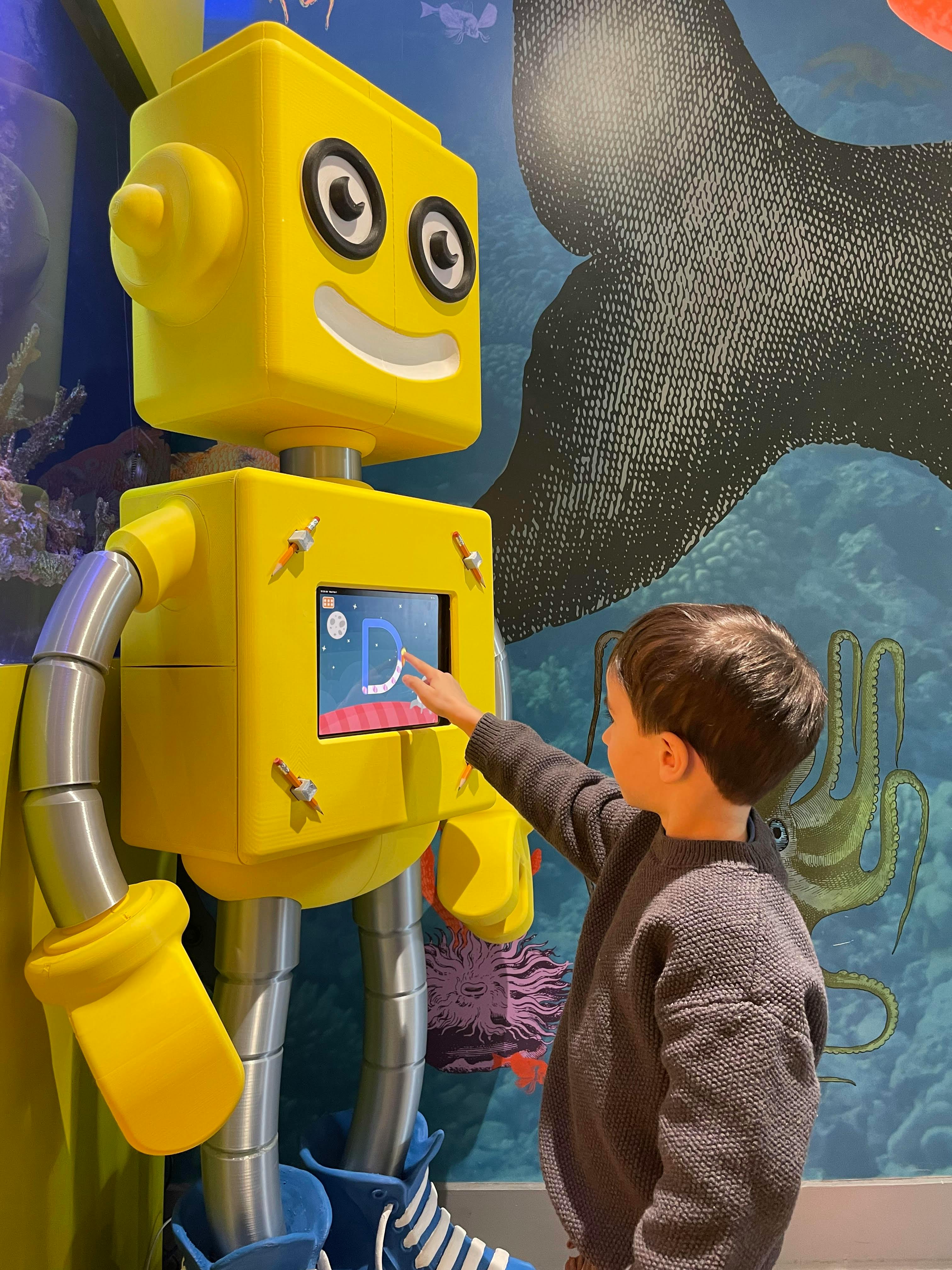 Child interacting with a yellow robot with a screen displaying the letter D