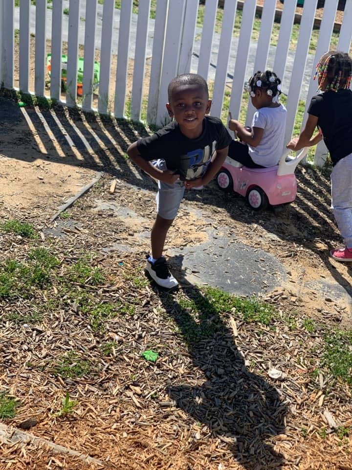 Child standing in front of a white fence with other children in the background