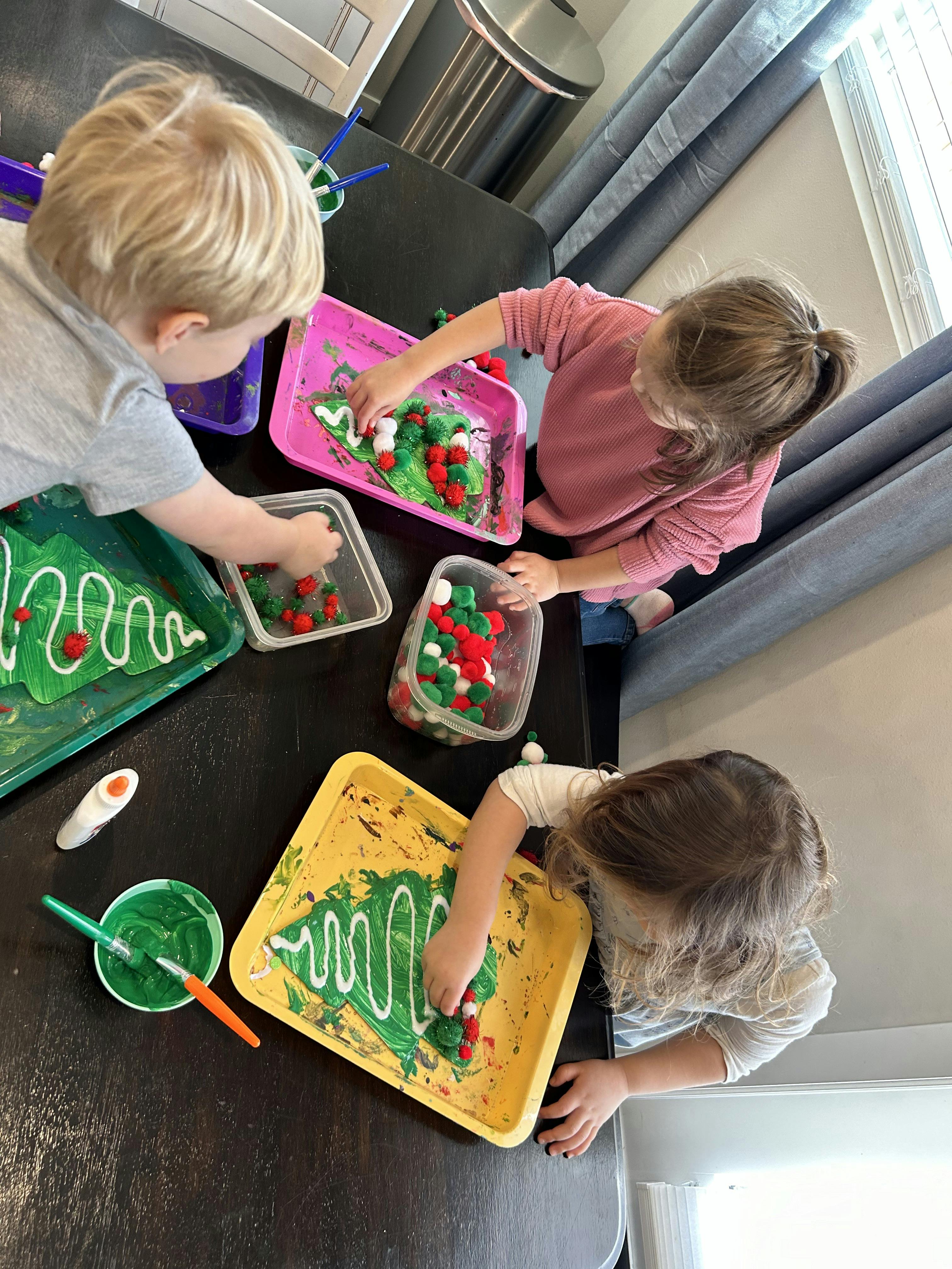 Three children decorating green tree shapes with pom-poms and white icing