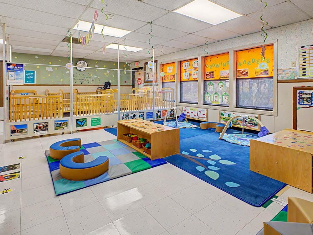 Cribs behind a partition in a brightly lit daycare room with playmats