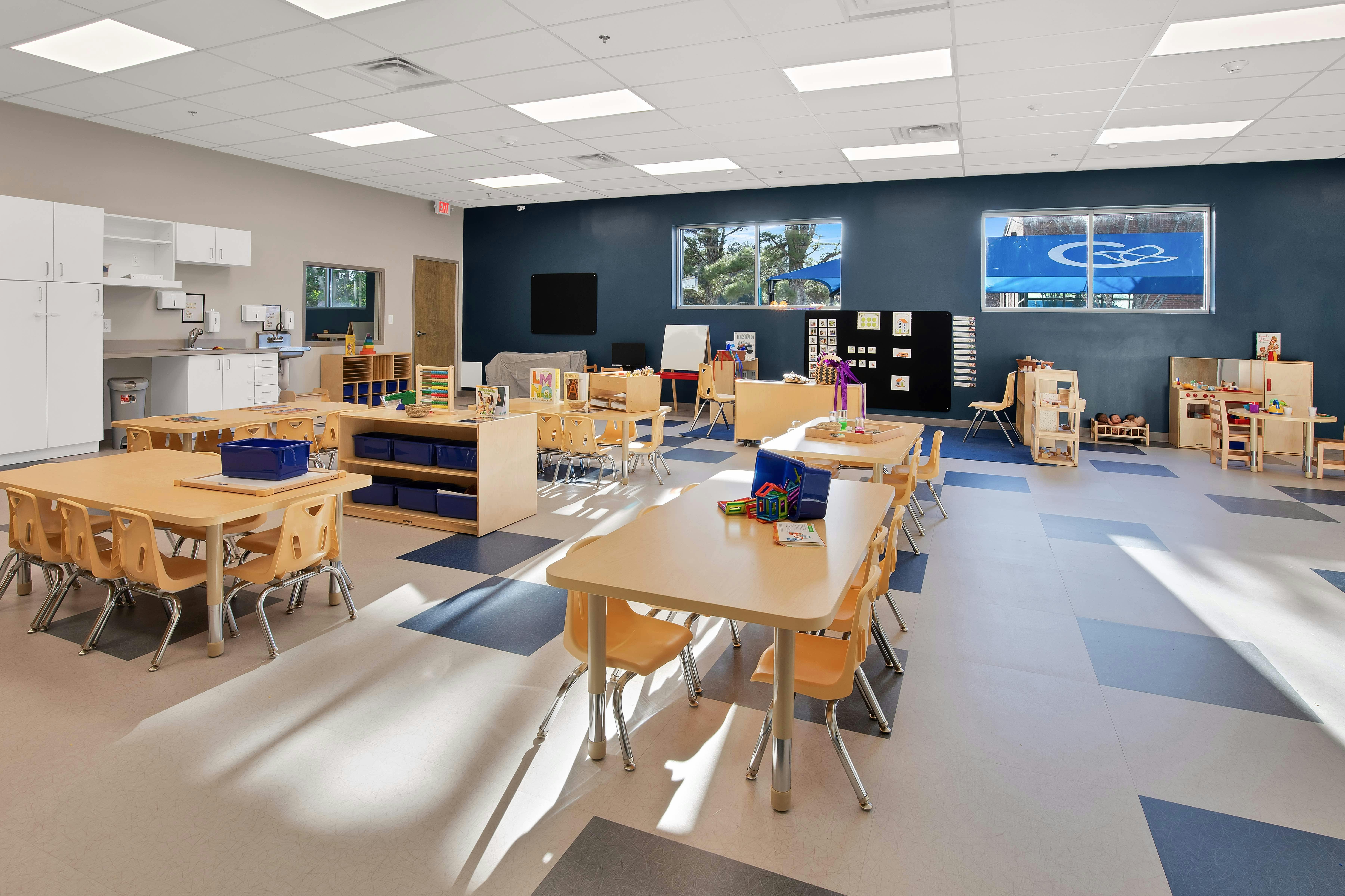 Empty preschool classroom with small tables and chairs, play kitchen, and storage cabinets
