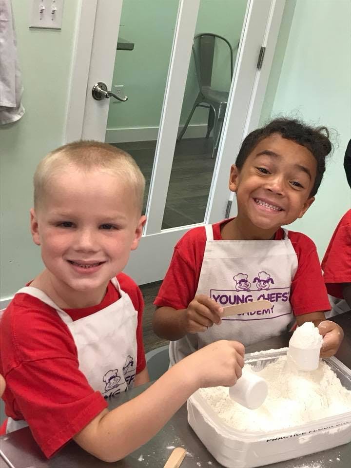 Two children in aprons with flour, one holding a measuring cup