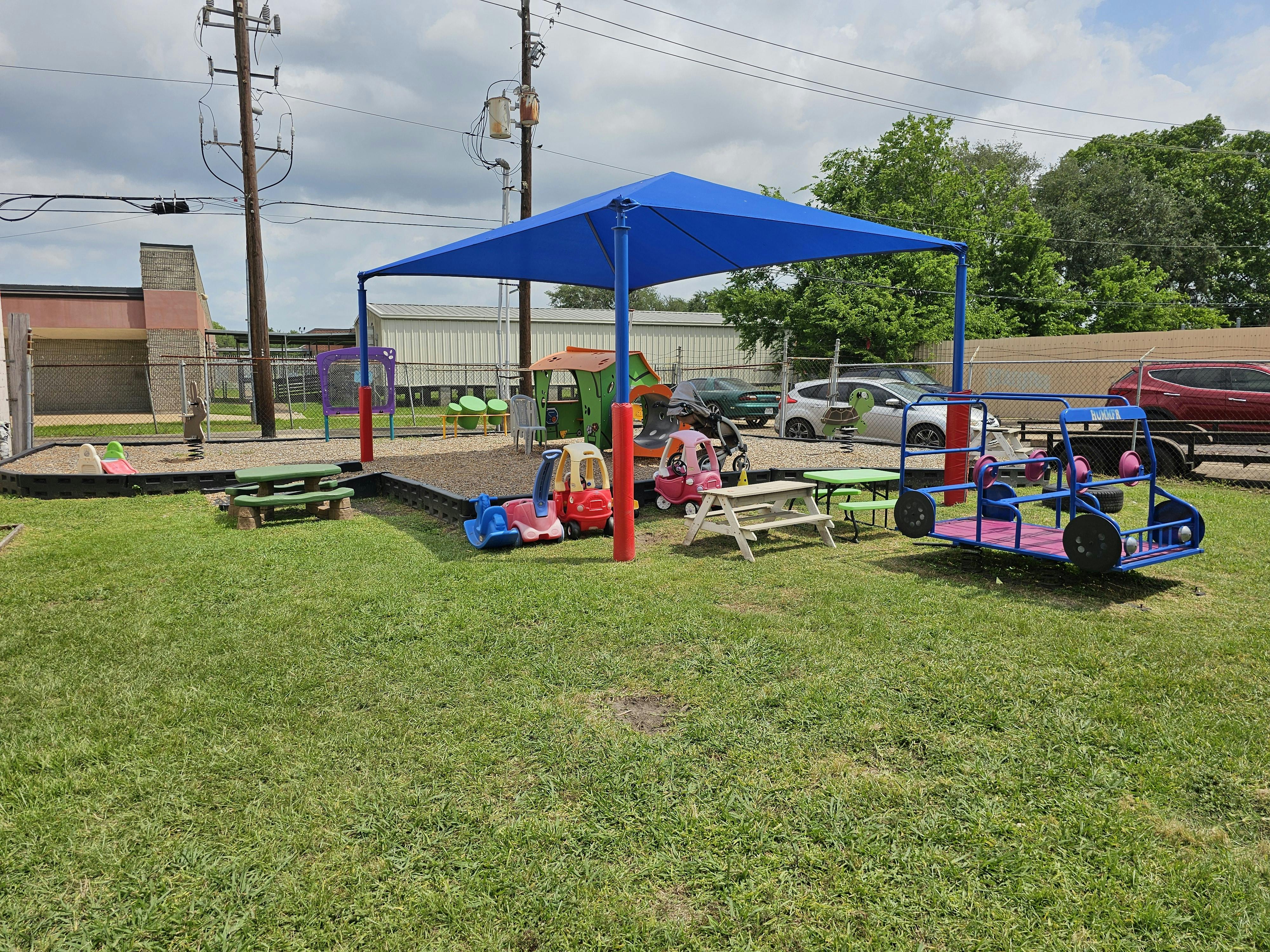 Outdoor play area with a blue canopy, picnic tables, and ride-on toys