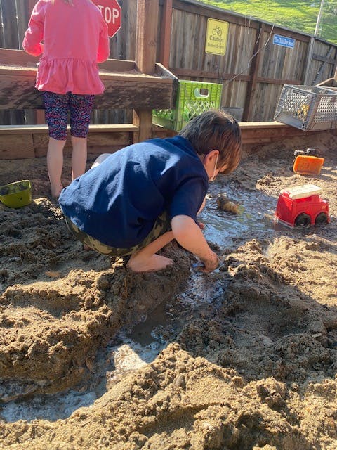Child playing in mud with toys, another child standing in background