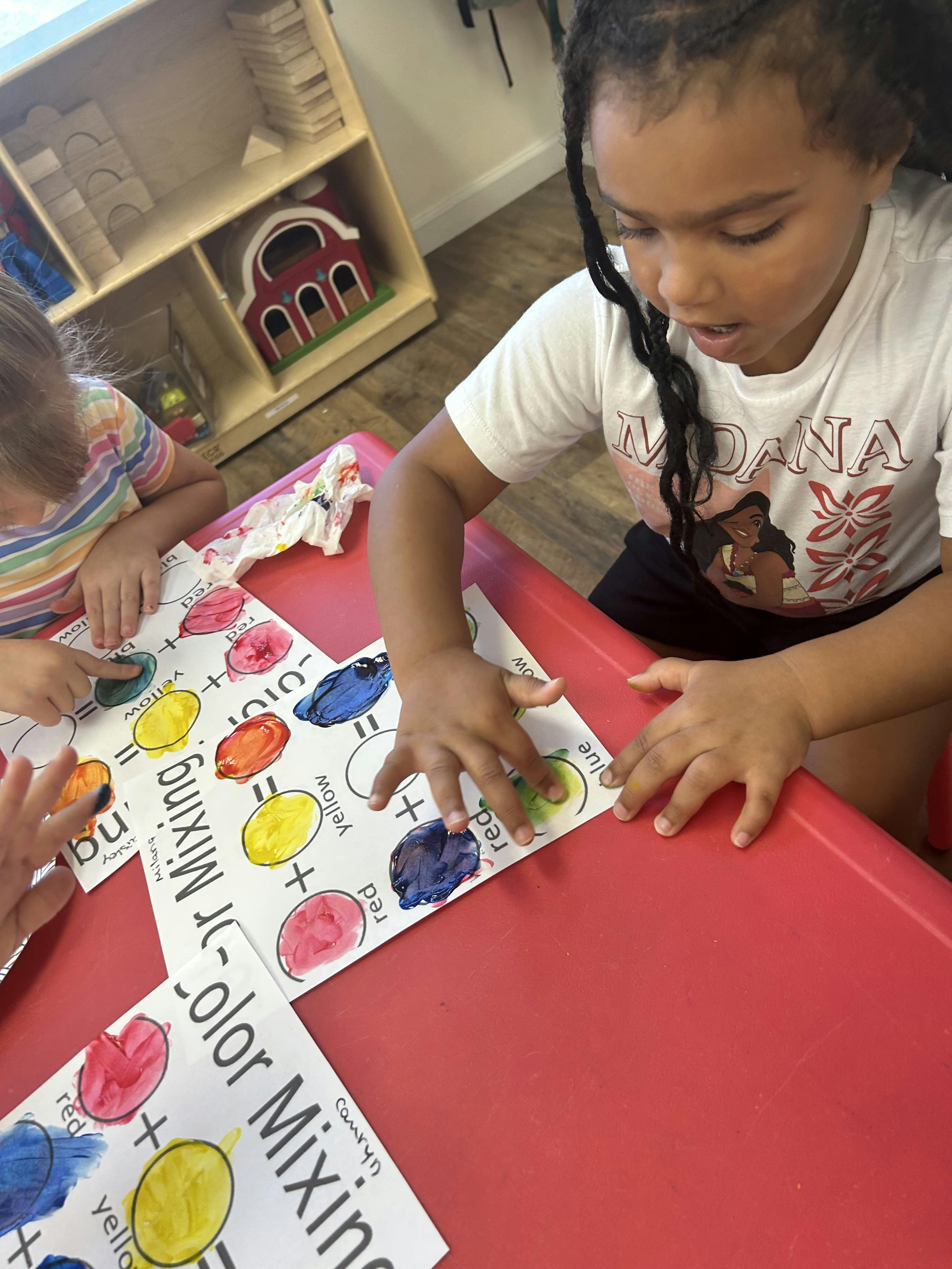 Children finger painting color mixing exercises on worksheets at a table