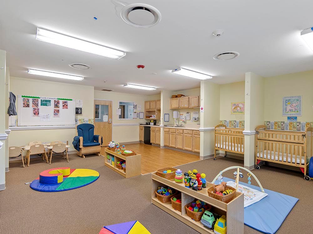 A colorful childcare room with cribs, play mats, and a small kitchen area