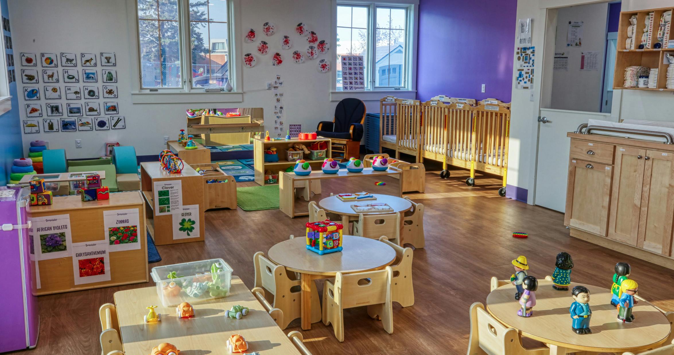 Classroom with small tables and chairs, cribs, and toys
