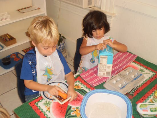 Two children at a table, one grating a carrot, the other opening a carton