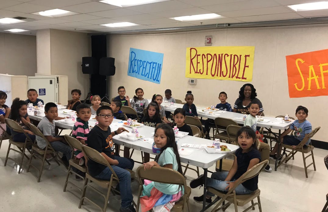 Children and adults sitting at tables in a room with signs that say "Respectful" and "Responsible"