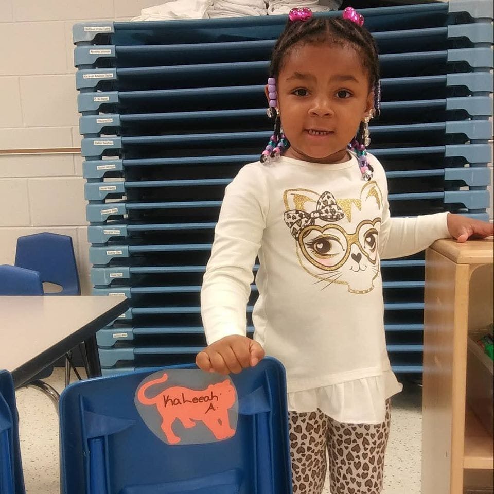 Child standing in a classroom, holding a blue chair