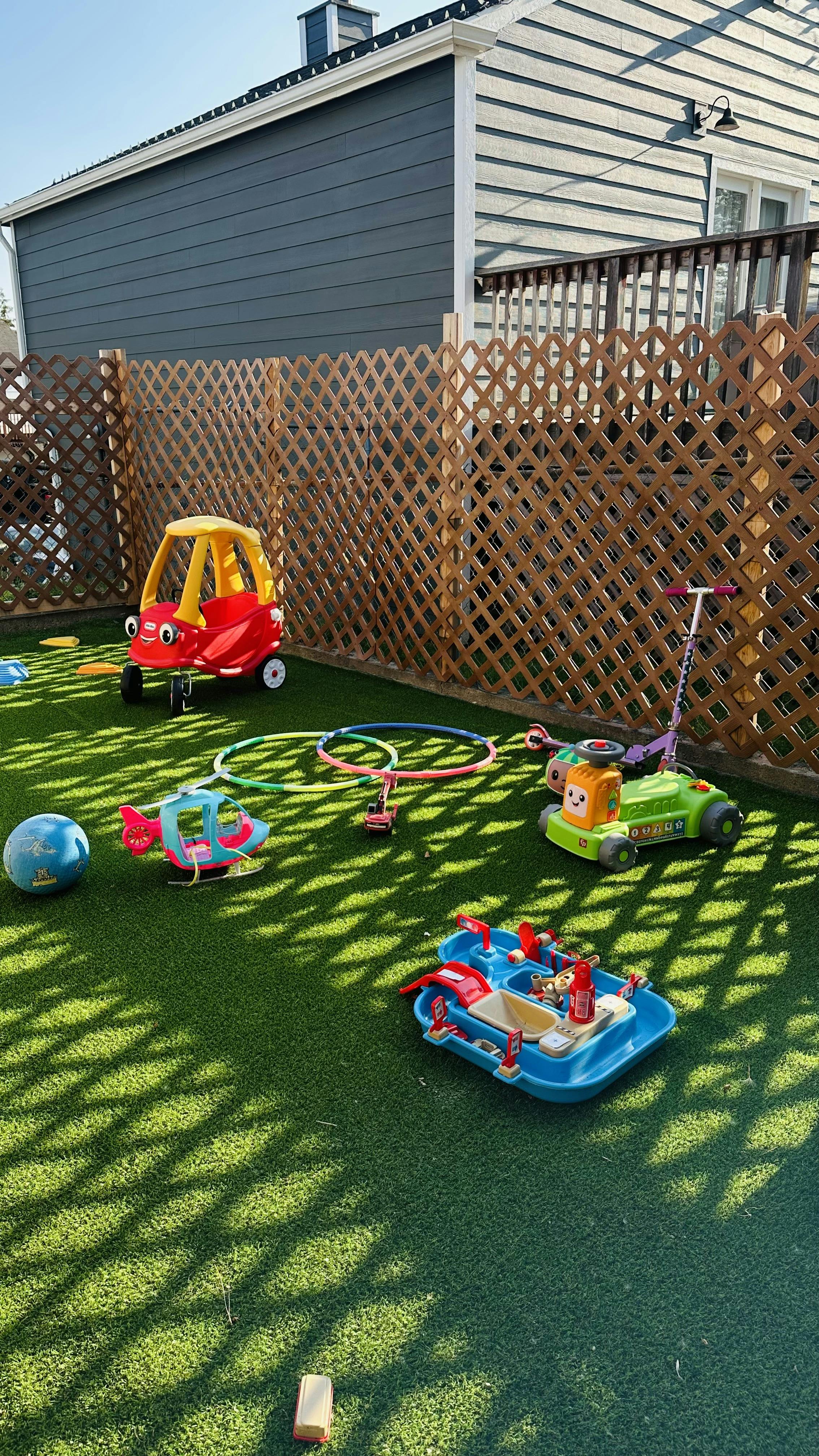 Various children toys scattered on artificial turf in a fenced backyard