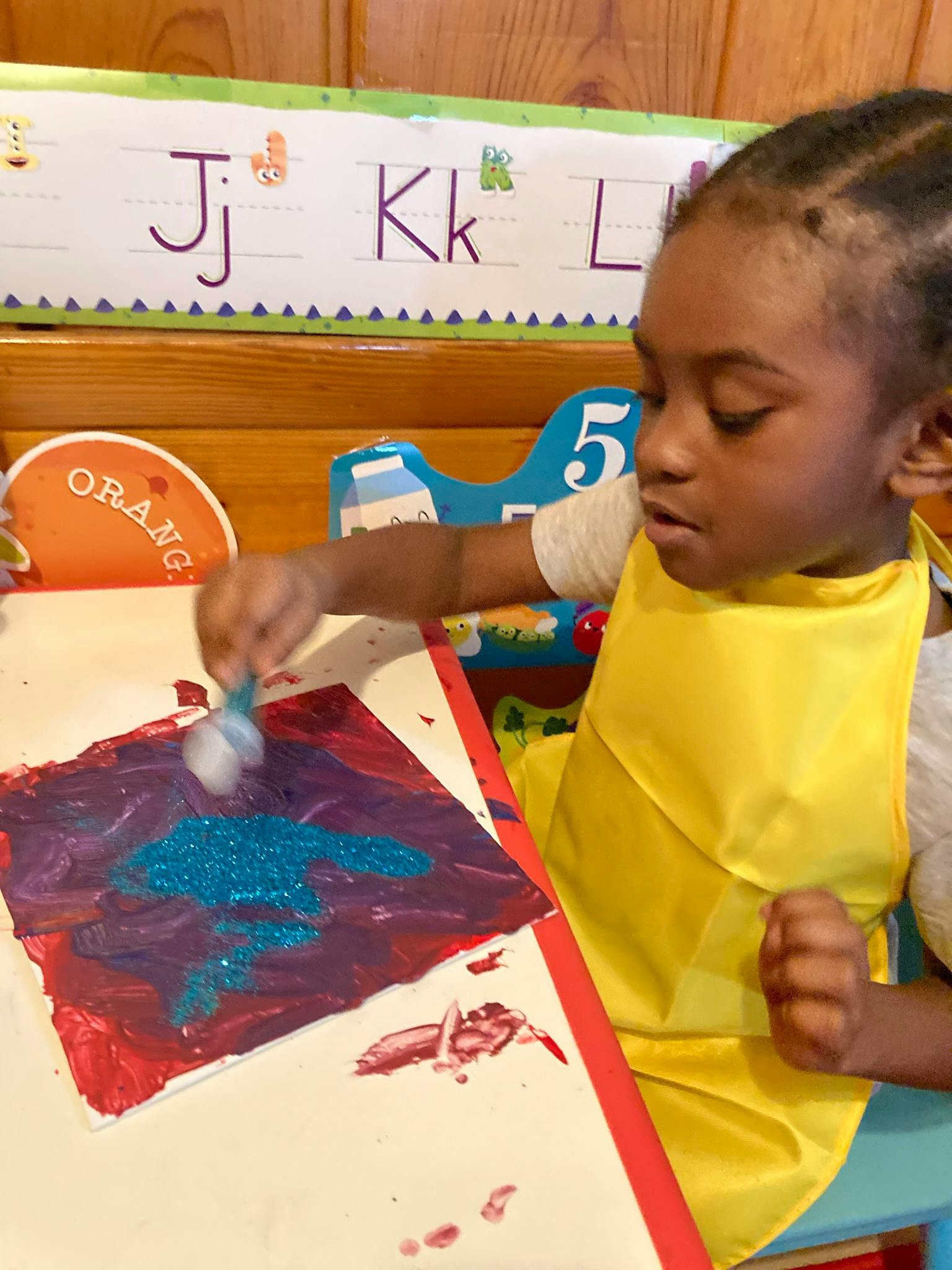 Child in a yellow bib painting with glitter on a red and purple canvas