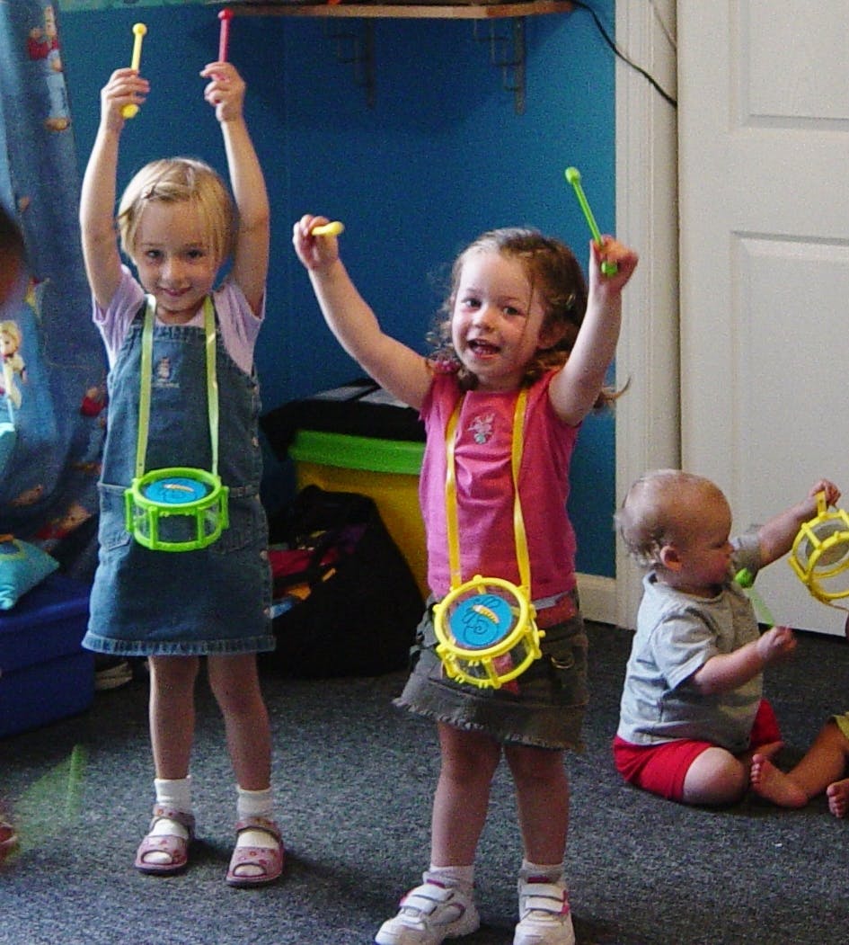 Two children holding drumsticks with toy drums around their necks, baby on floor