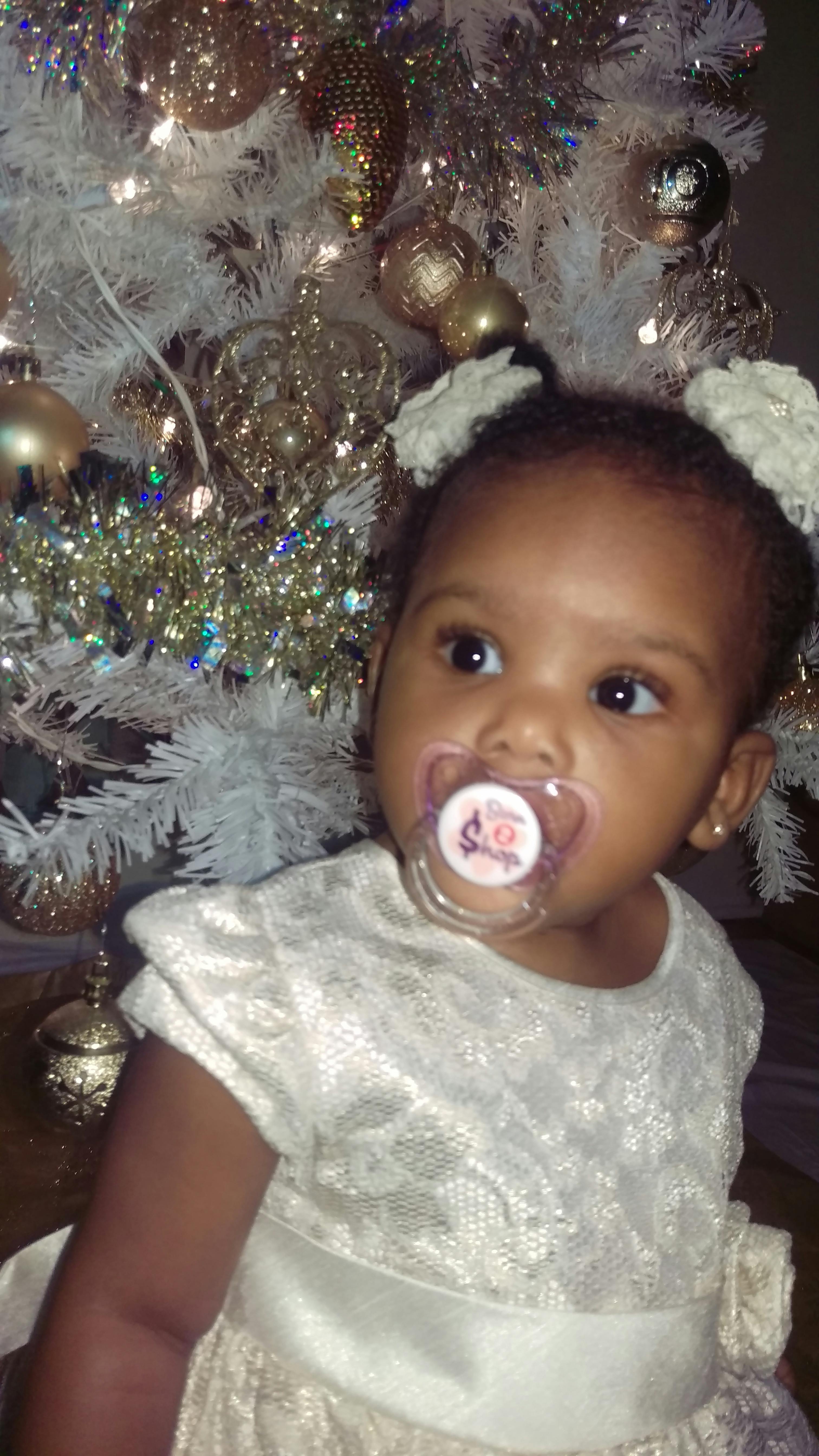 Child with pacifier wearing a light-colored dress in front of a Christmas tree