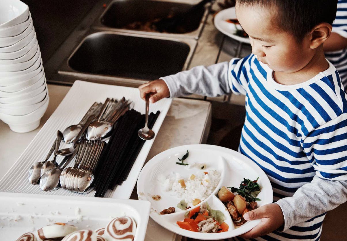 Child holding a divided plate with food and a spoon over a serving tray