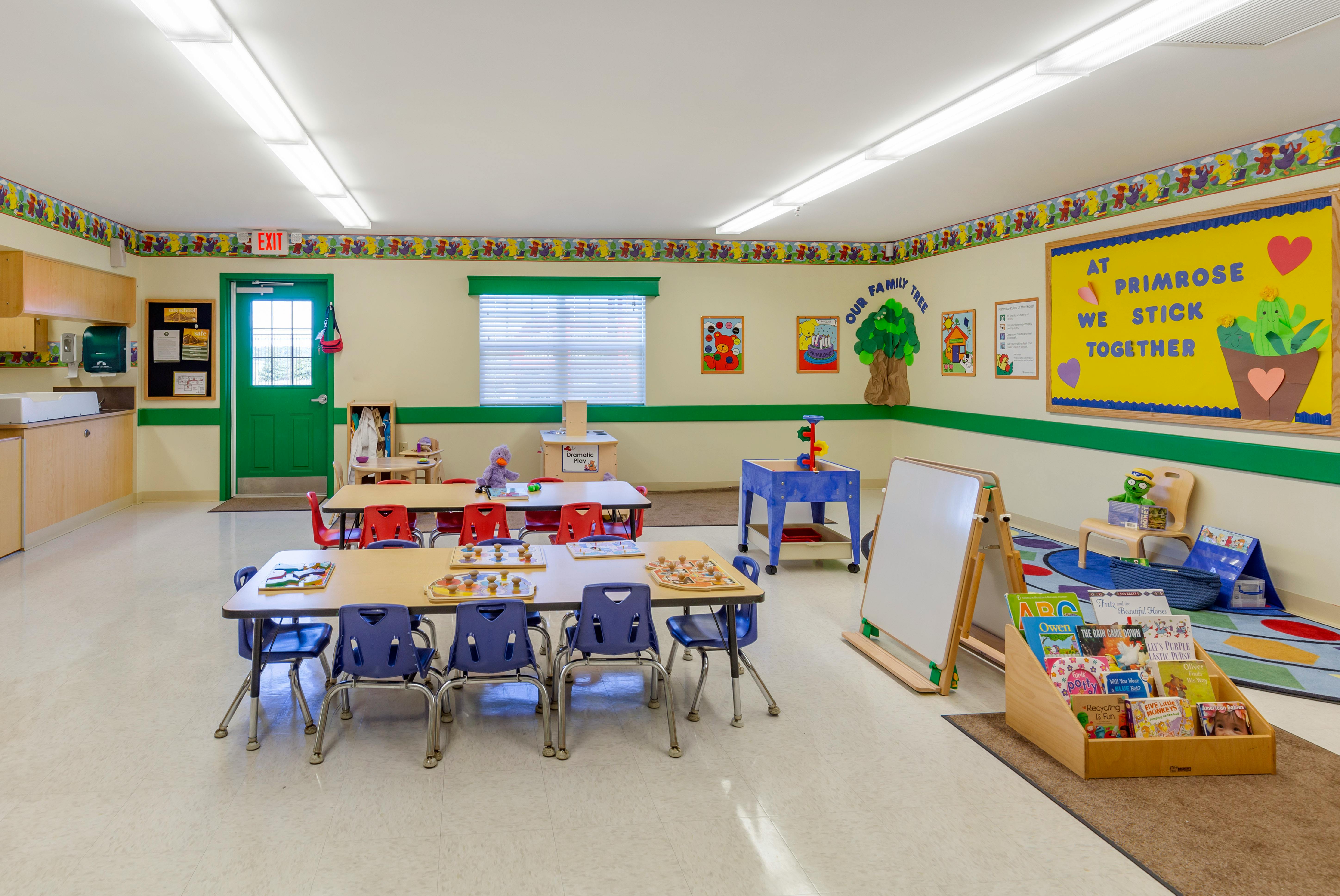 Classroom with child-sized tables and chairs, a play area, and a bulletin board that reads "At Primrose We Stick Together"