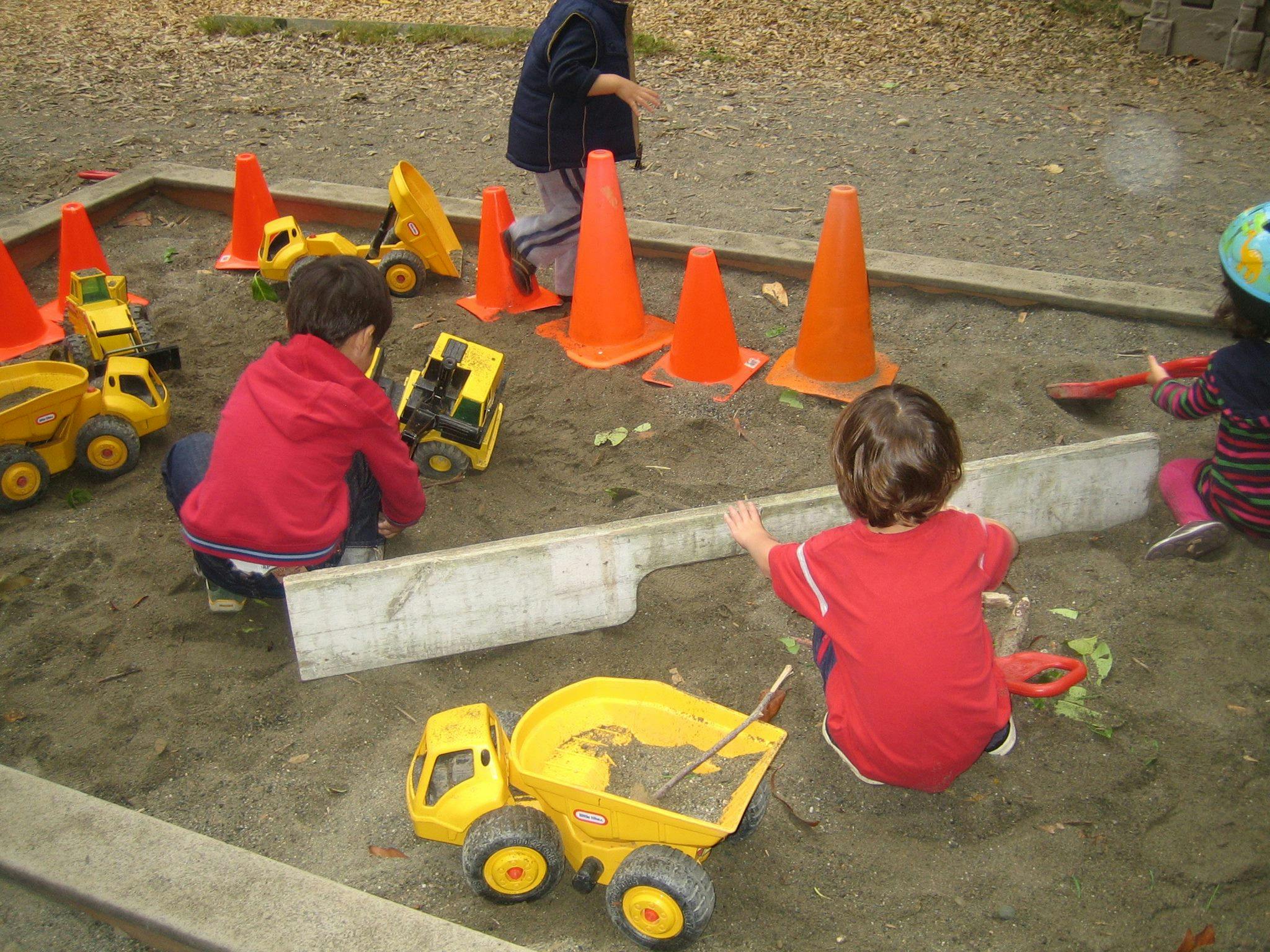 Children playing in a sandbox with toy trucks and orange cones