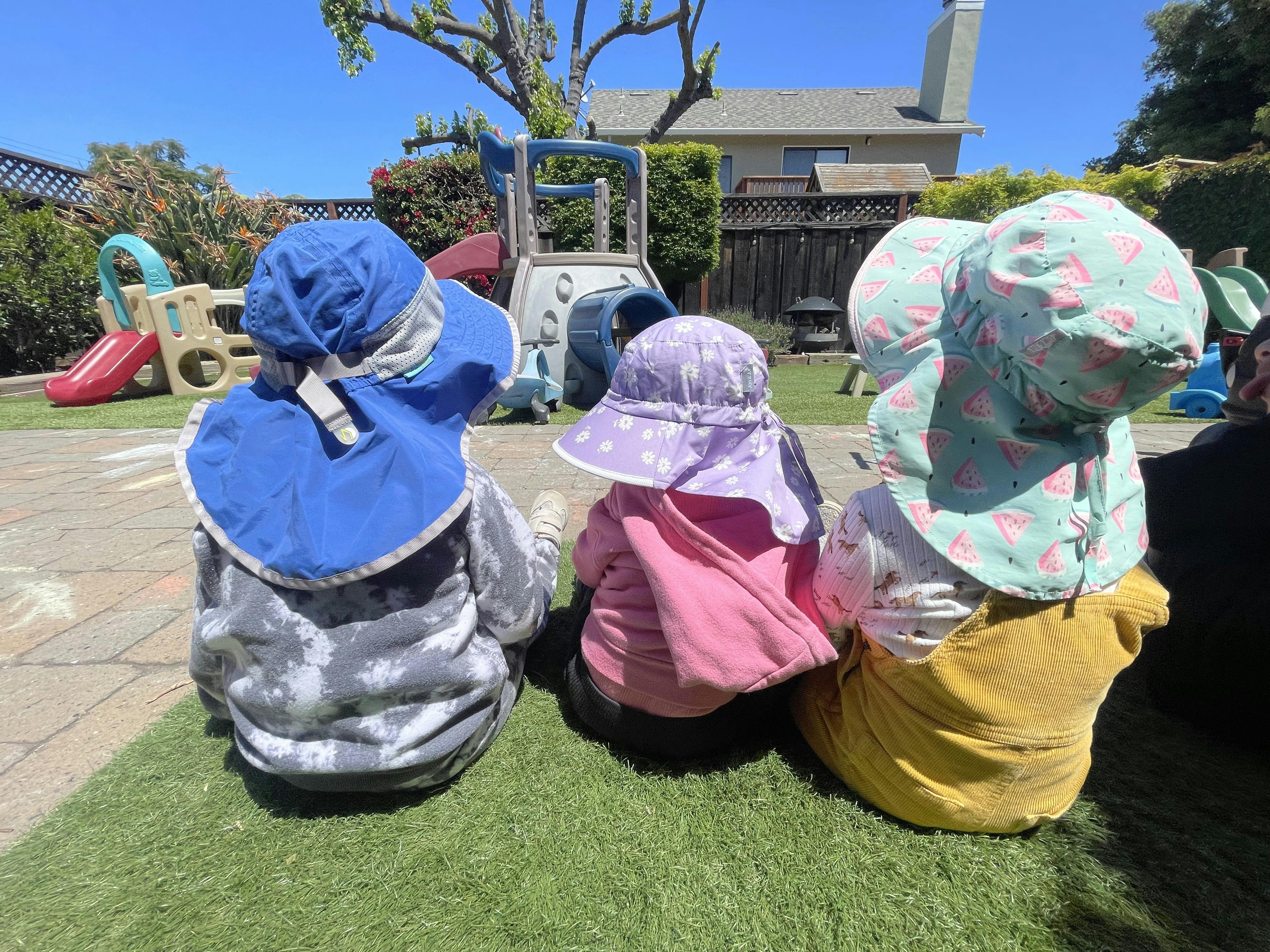 Three children in sun hats sitting on artificial grass, looking at a playground