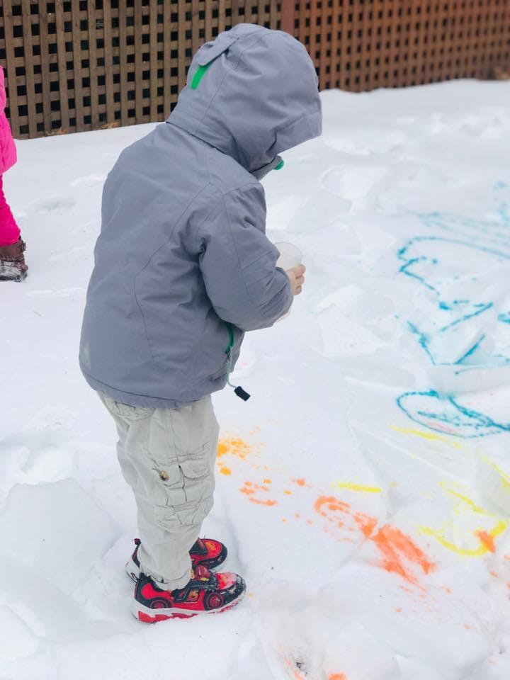 Child in winter coat holding a cup and looking at colorful drawings in snow