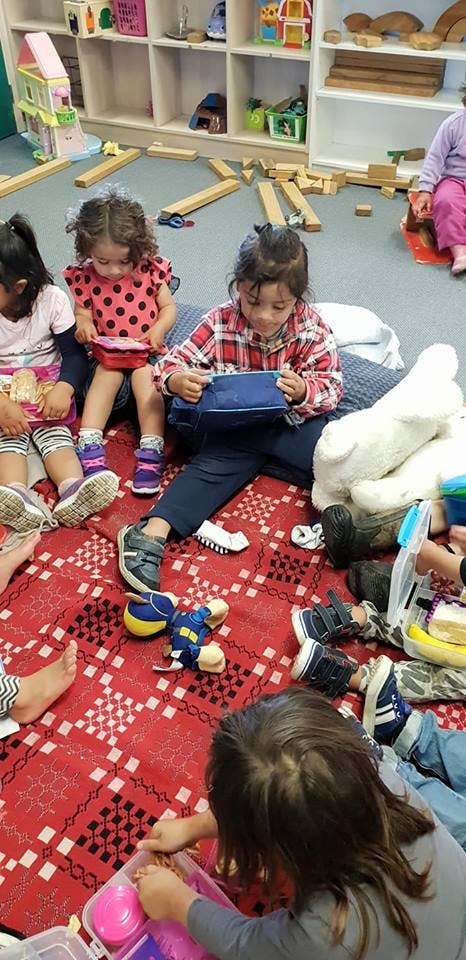 Children sitting on a red patterned rug, some holding lunchboxes