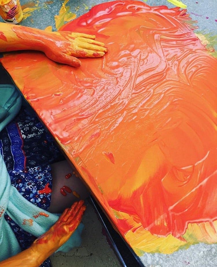 Person's hands covered in paint on a canvas with orange and yellow paint