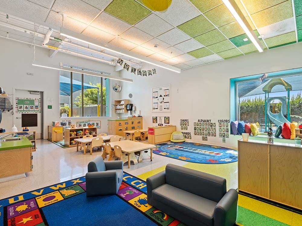 Colorful classroom with small tables and chairs, play mats, and a slide visible through a window
