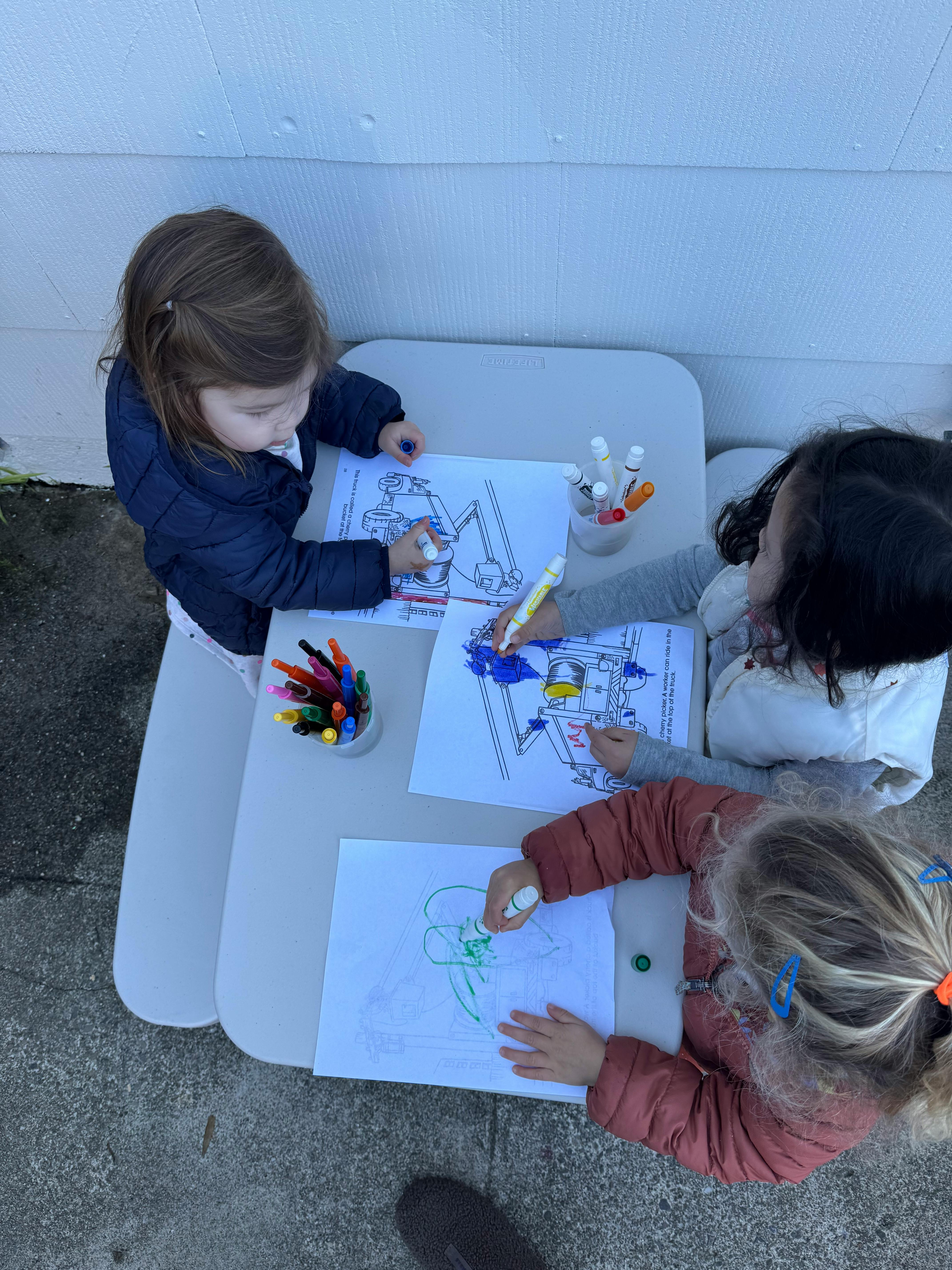 Three children coloring at a small table outdoors