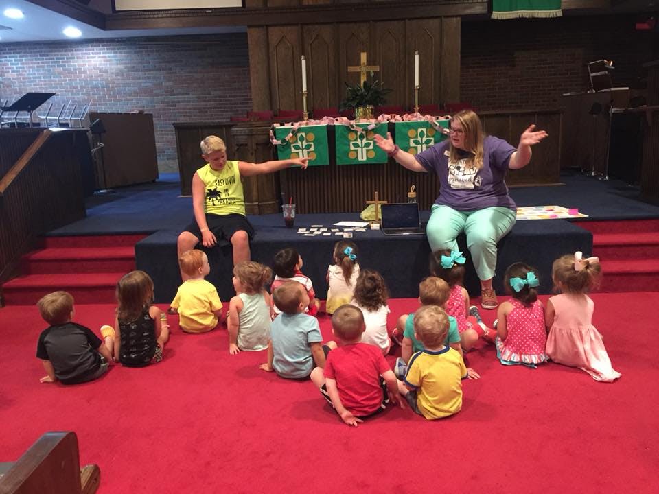 Two adults interacting with a group of children sitting on a red floor