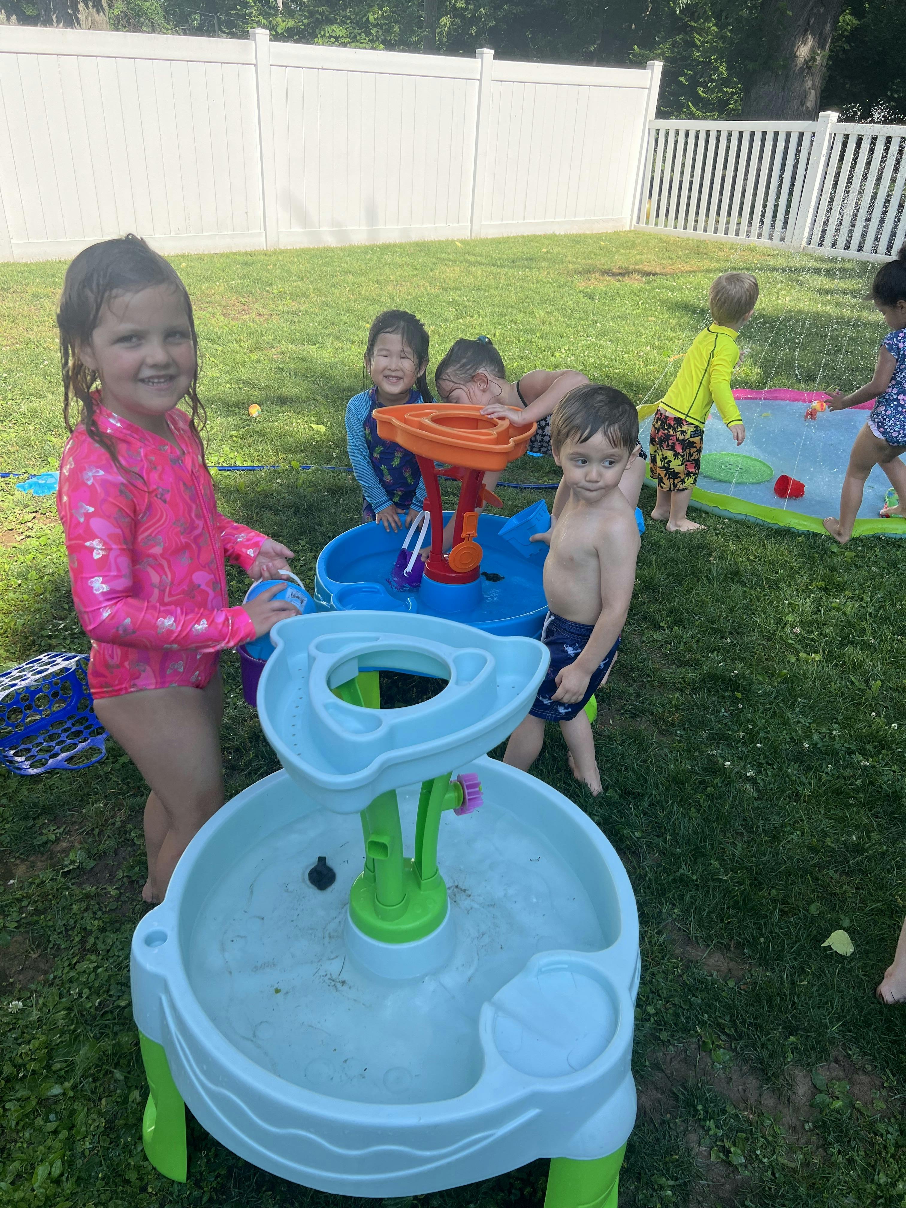 Group of children playing with water tables and sprinklers in a grassy backyard