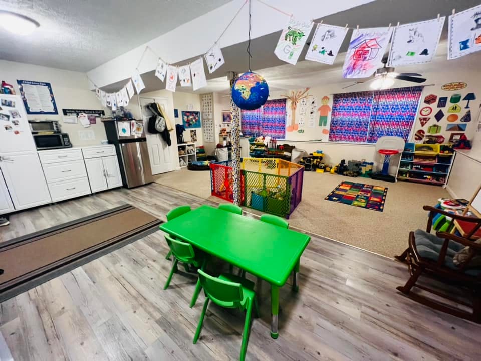 A classroom with a green table and chairs, kitchen cabinets, and children's artwork