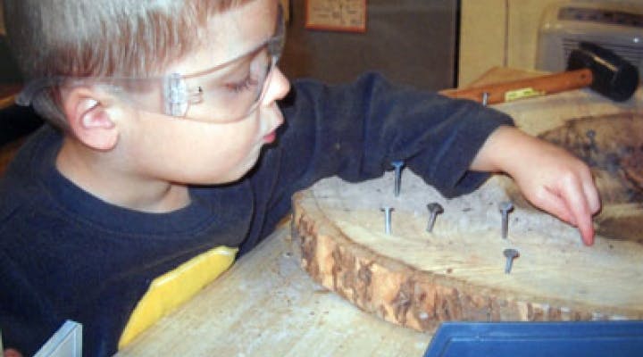 Child in safety glasses hammering nails into a log slice