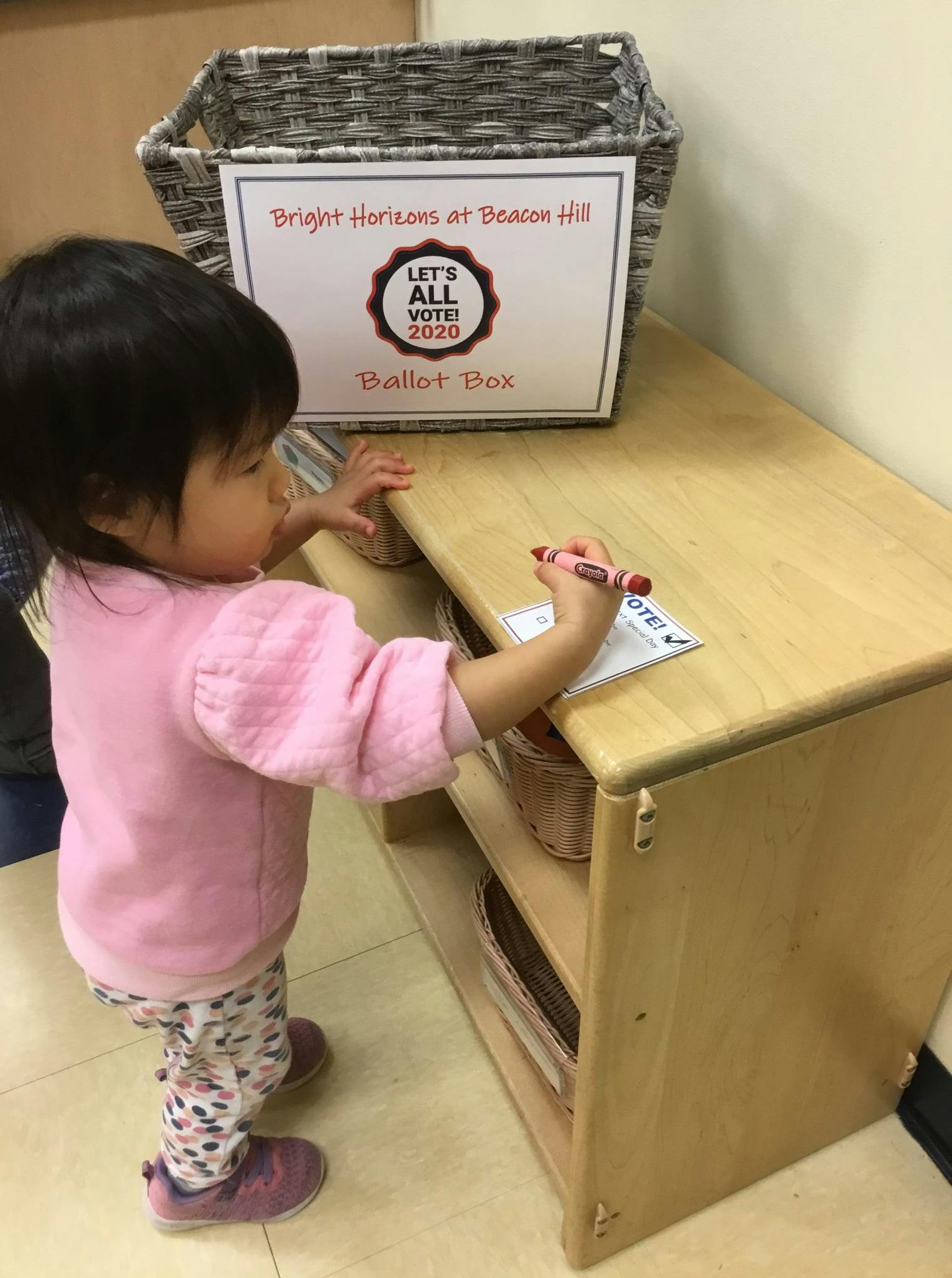Child writing on paper with a crayon next to a ballot box sign